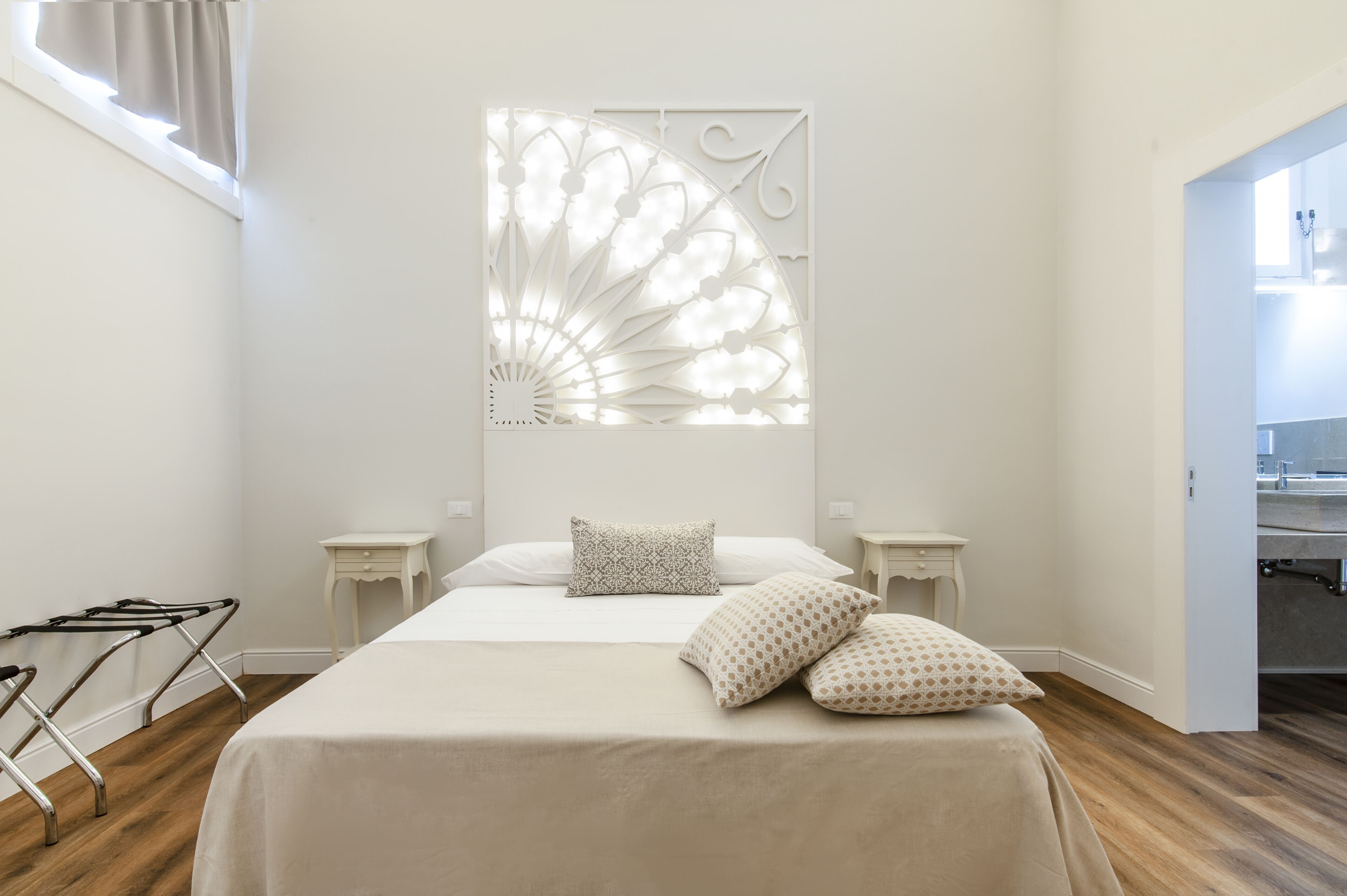 A bedroom with a bed, two bedside tables, a window with curtains, a wall-mounted decorative panel, and a wooden floor.