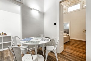 Una habitación moderna con una mesa redonda, sillas blancas, suelo de madera y una cama al fondo.
