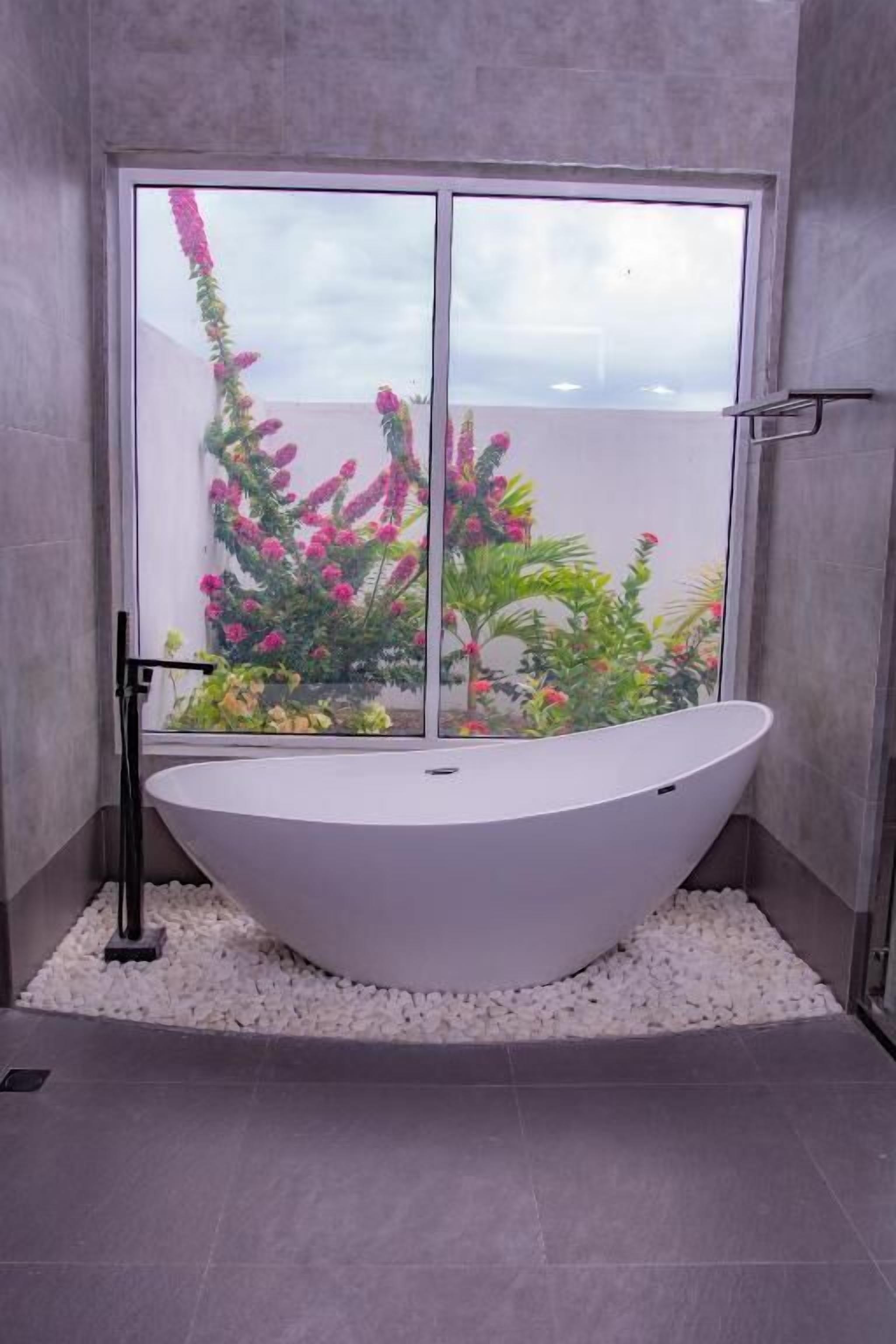 A modern bathroom with a freestanding bathtub, a glass shower enclosure, and a view of a garden with pink flowers.