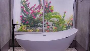 A modern bathroom with a freestanding bathtub, a glass shower enclosure, and a view of a garden with pink flowers.