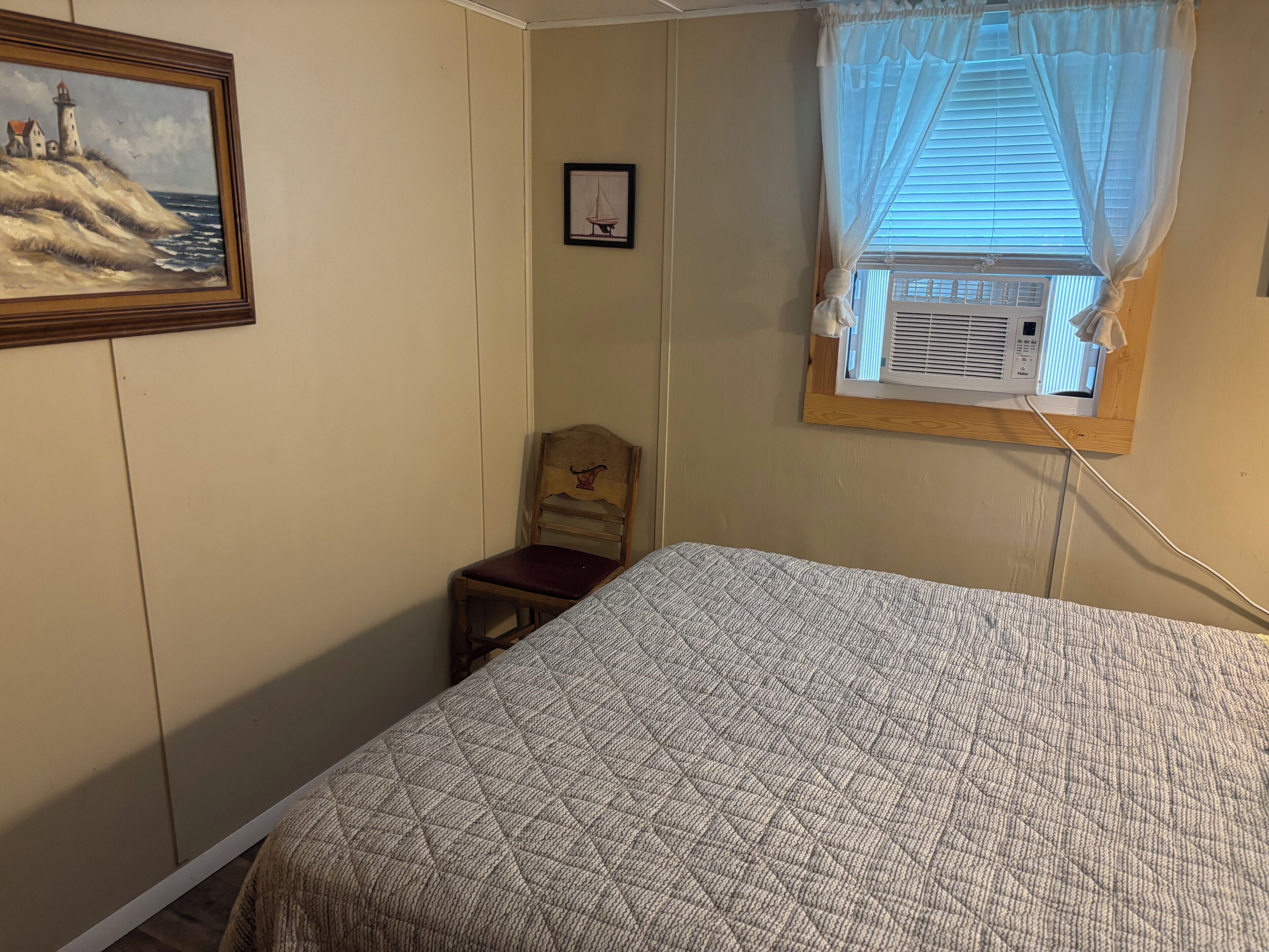 A bedroom with a bed, a chair, a painting of a lighthouse, and a window with an air conditioning unit.