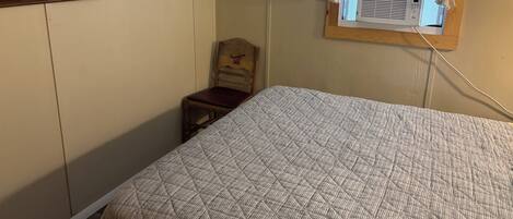 A bedroom with a bed, a chair, a painting of a lighthouse, and a window with an air conditioning unit.