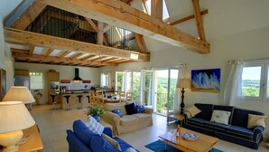 A spacious living area with a high ceiling, wooden beams, a kitchen in the background, and a large window offering a view of the outdoors. - Beautifully Finished House, Modern Interiors, Private Heated Pool & Fabulous Location near Martel! (Martel)