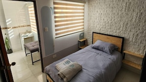 Quarto com cama, mesa de cabeceira, janela com persianas, parede com textura de pedra e porta que dá para o banheiro.