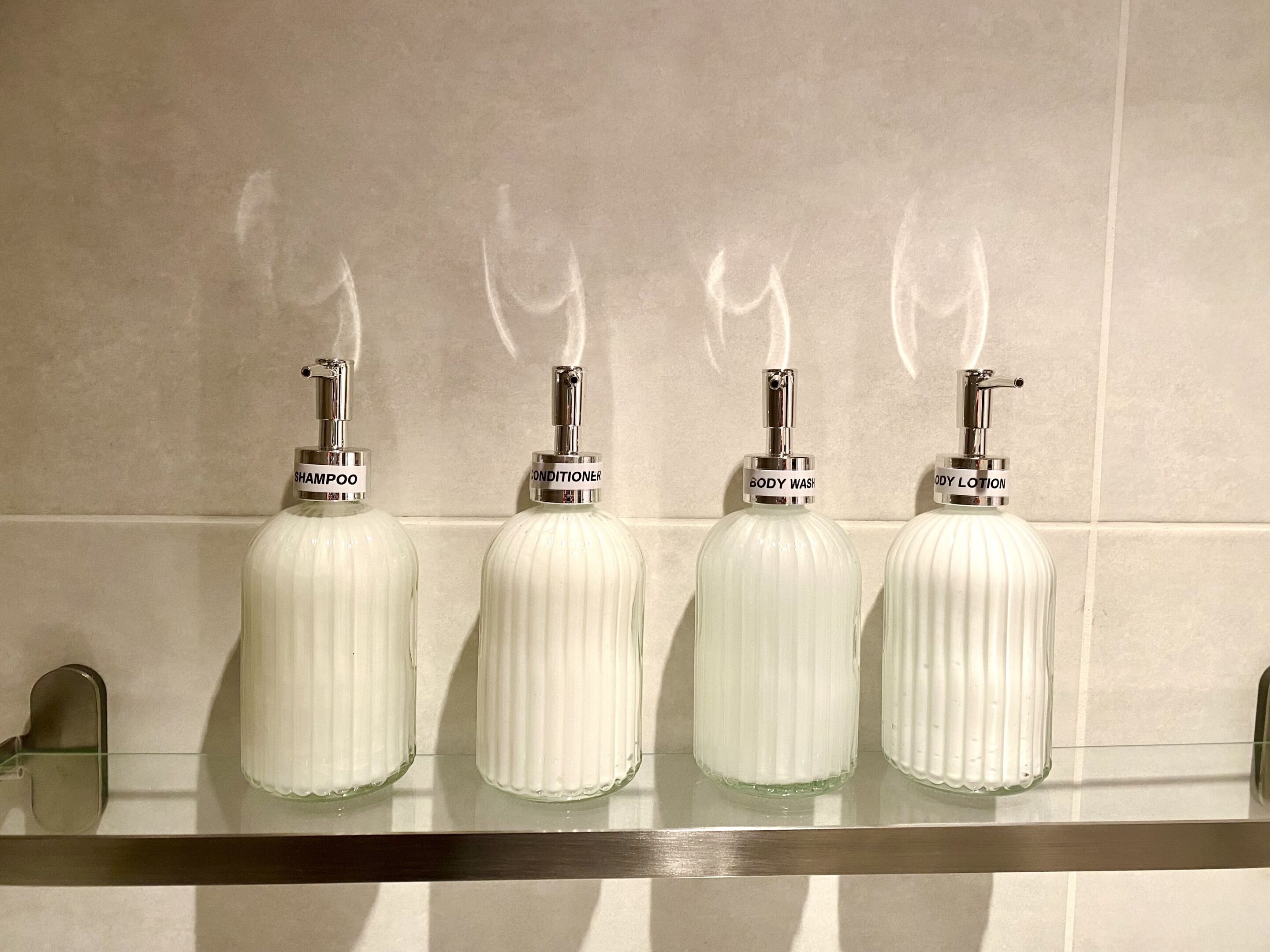 Four white bottles with labels on a shelf against a tiled wall.