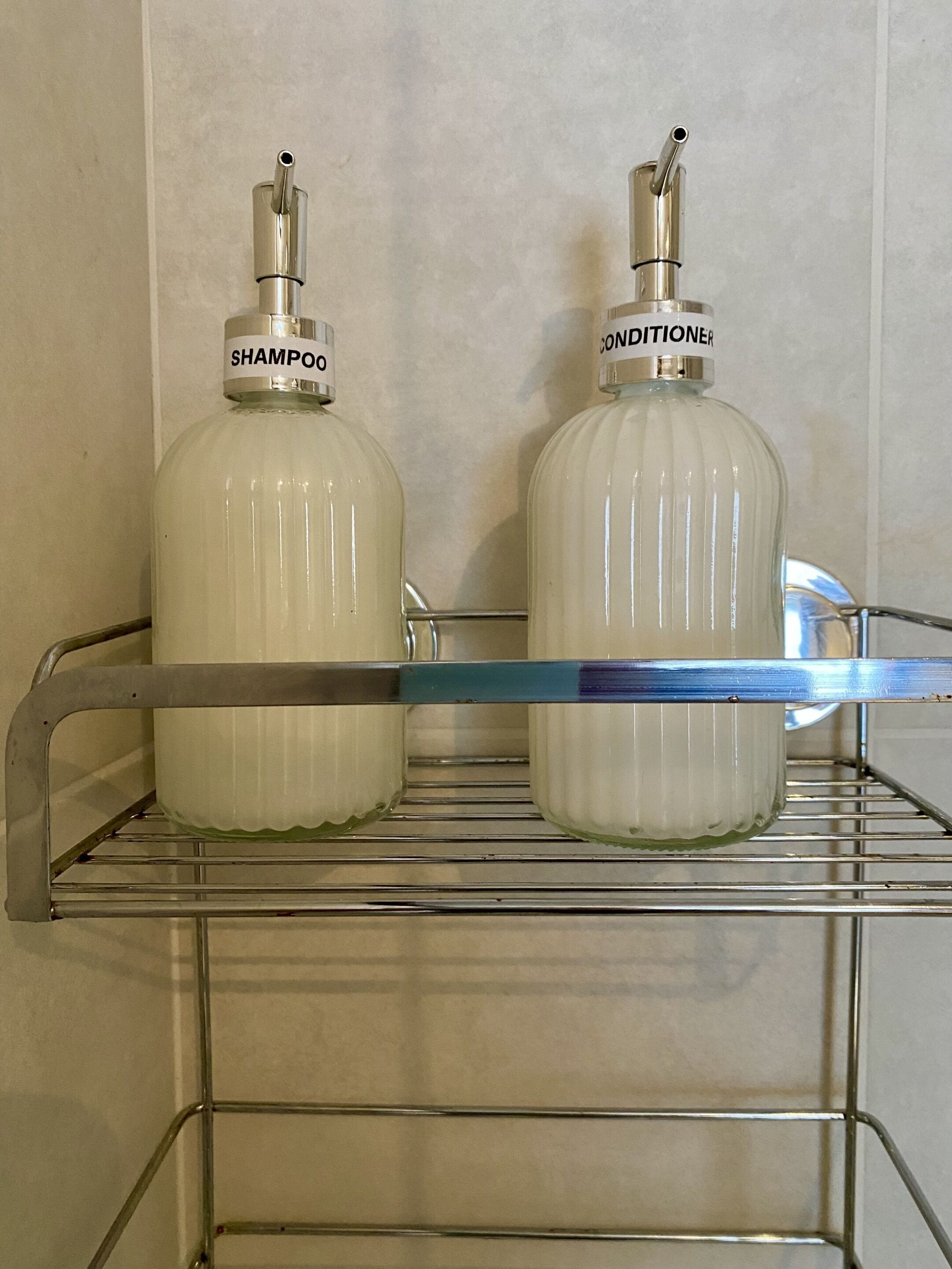 Dos botellas grandes de champú y acondicionador sobre una estantería metálica en un baño.