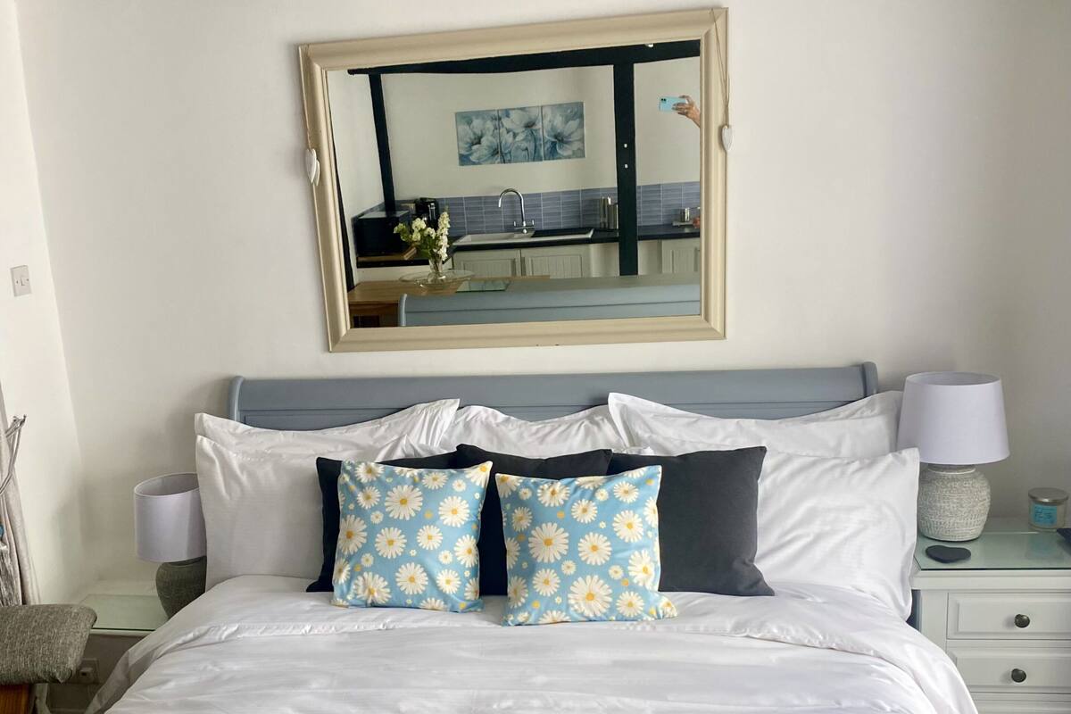 A neatly made bed with white and blue floral pillows, a grey headboard, and a bedside table with a lamp and a small decorative item.