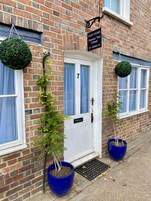 A brick building with a white door labeled "7" and a sign indicating "Guest Rooms Chestnut Tree Cottage." There are two potted plants on the ground, and a hanging spherical decoration above the door.