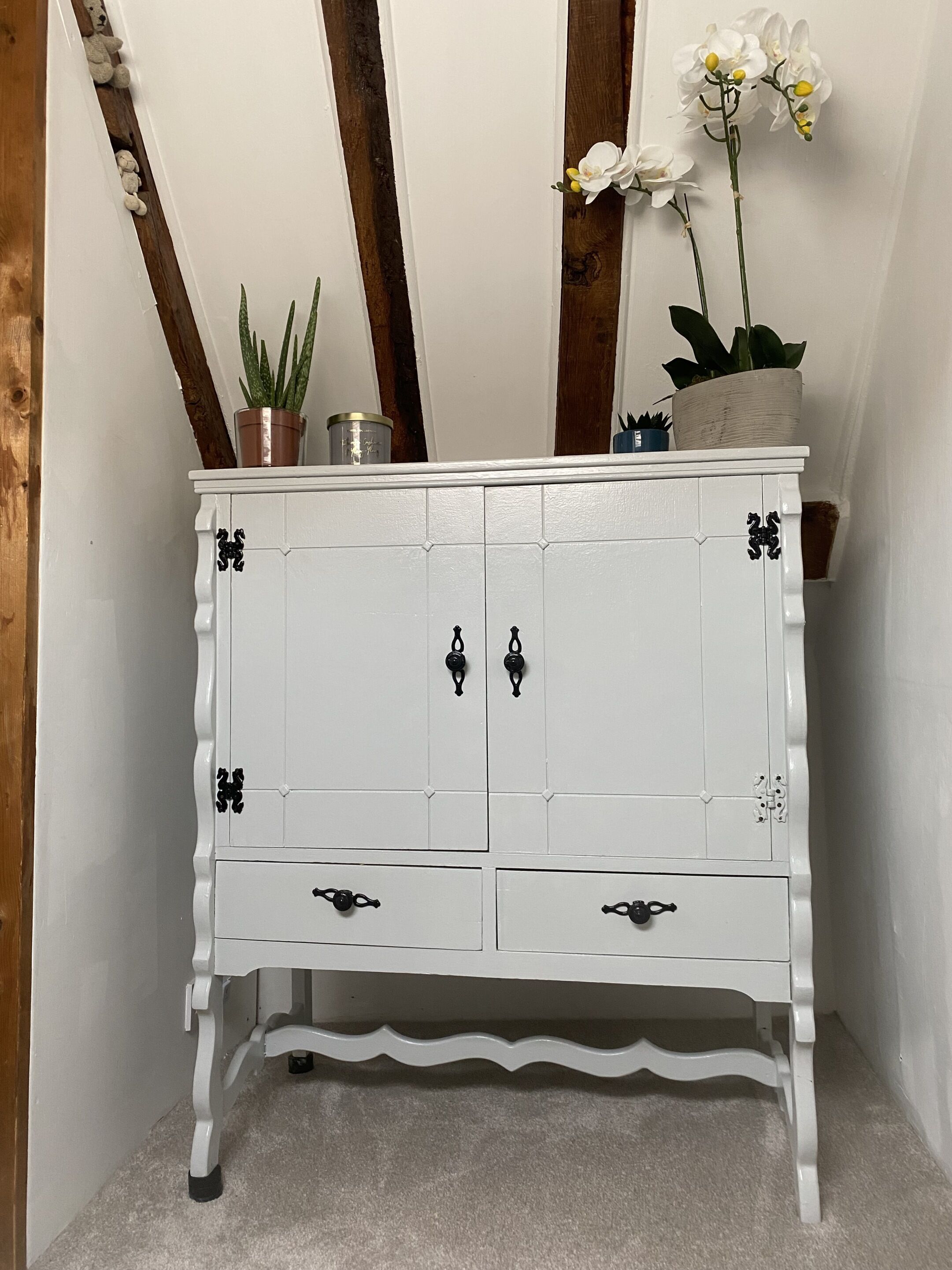 Un mueble blanco con manijas negras y detalles decorativos, situado frente a una viga de madera y una pared blanca.