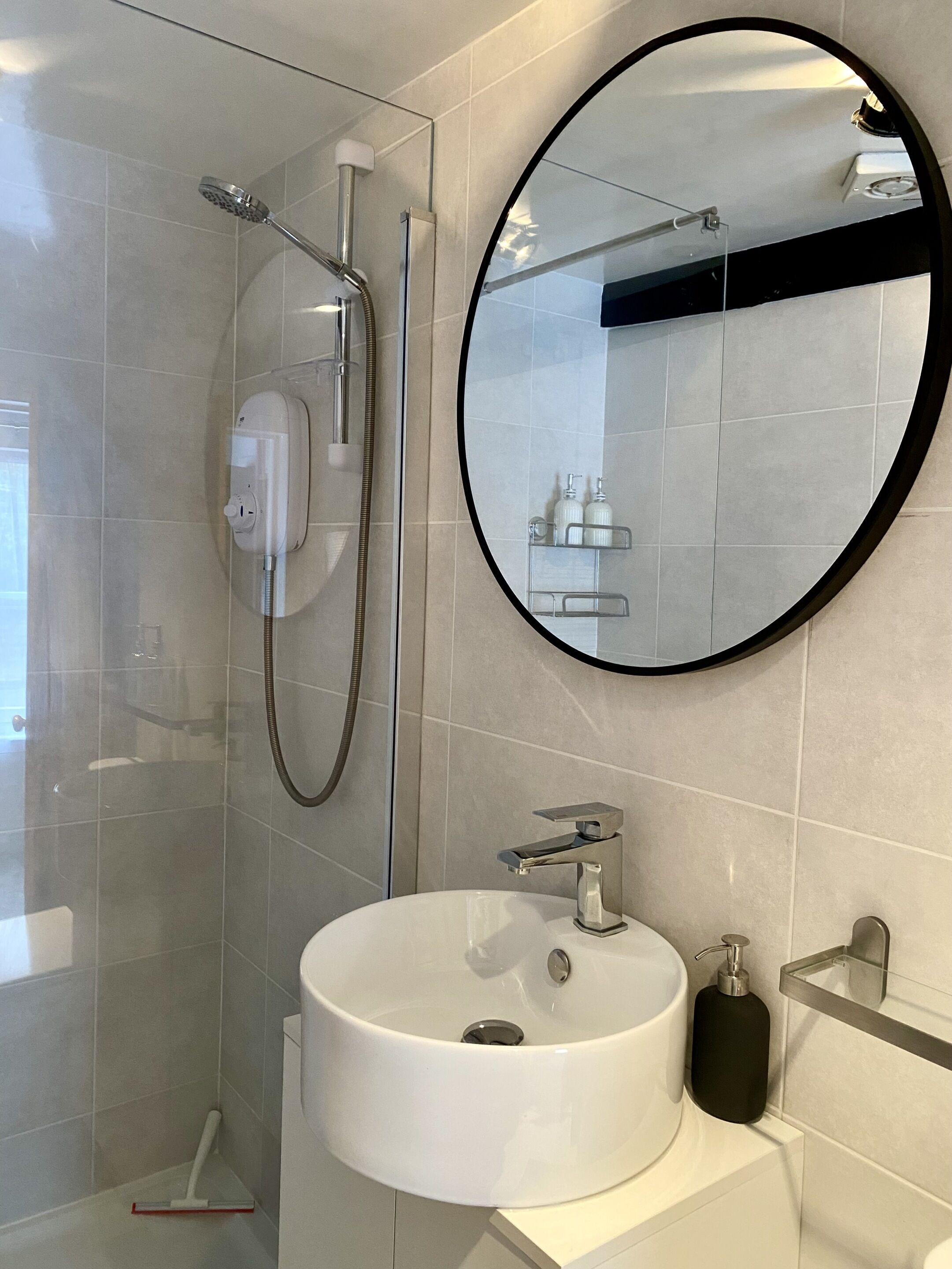 A modern bathroom with a glass-enclosed shower, a round mirror, and a white sink.