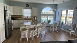 A spacious kitchen with white cabinets, a central island, and a dining area with wicker chairs. - Waterfront 4 BR retreat at the sound side and only 100 steps to the ocean beach. (Emerald Isle)
