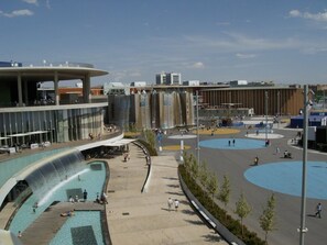 Une place urbaine moderne agrémentée d’un élément aquatique, de zones d’assise et d’un grand bâtiment doté d’un toit incurvé.