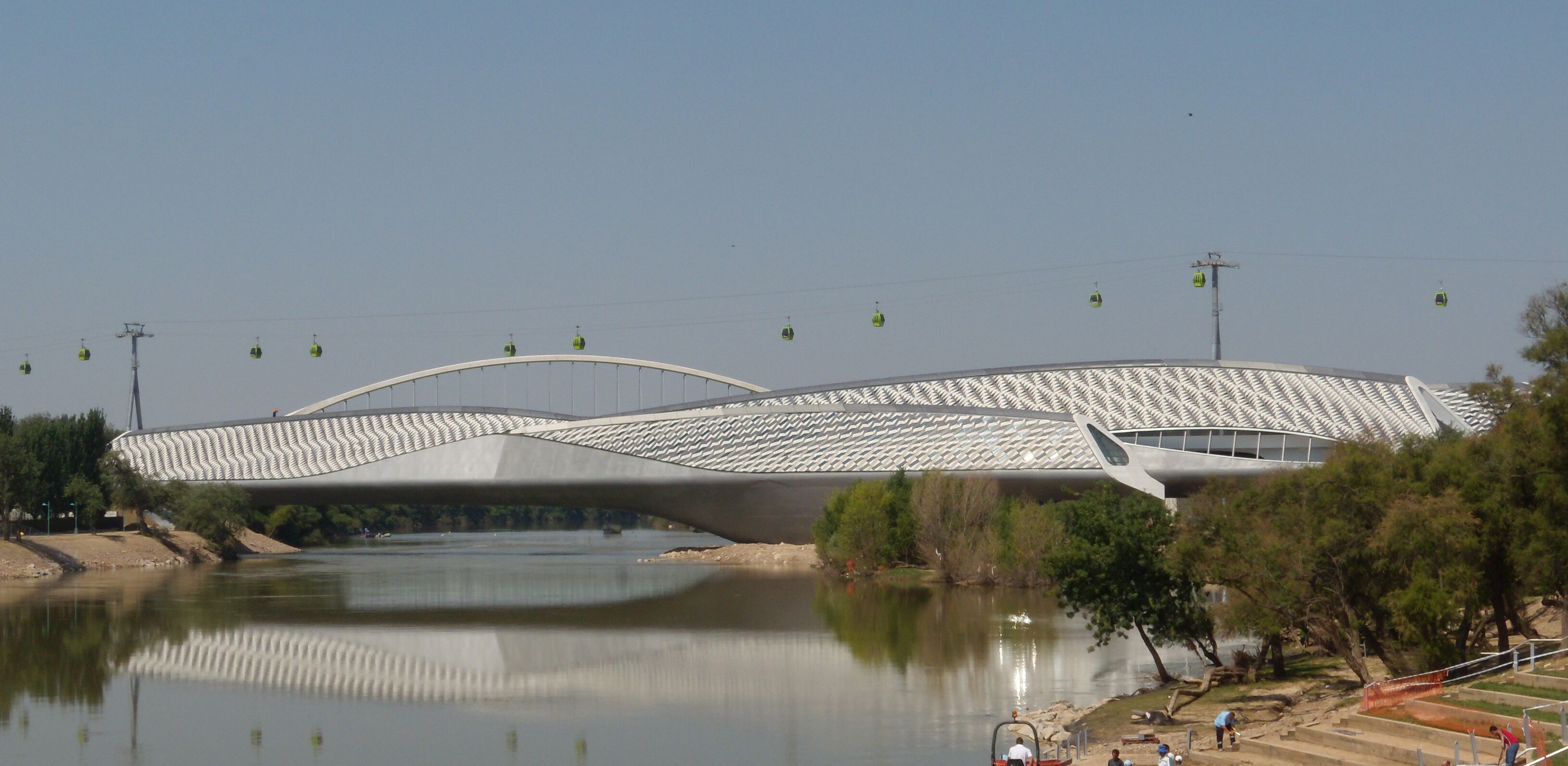 A modern architectural bridge with a unique wavy design, over a calm river, with cable cars visible in the background.