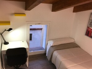 Une petite pièce comprenant un bureau, une chaise et un lit. Il y a une fenêtre donnant sur l’extérieur.