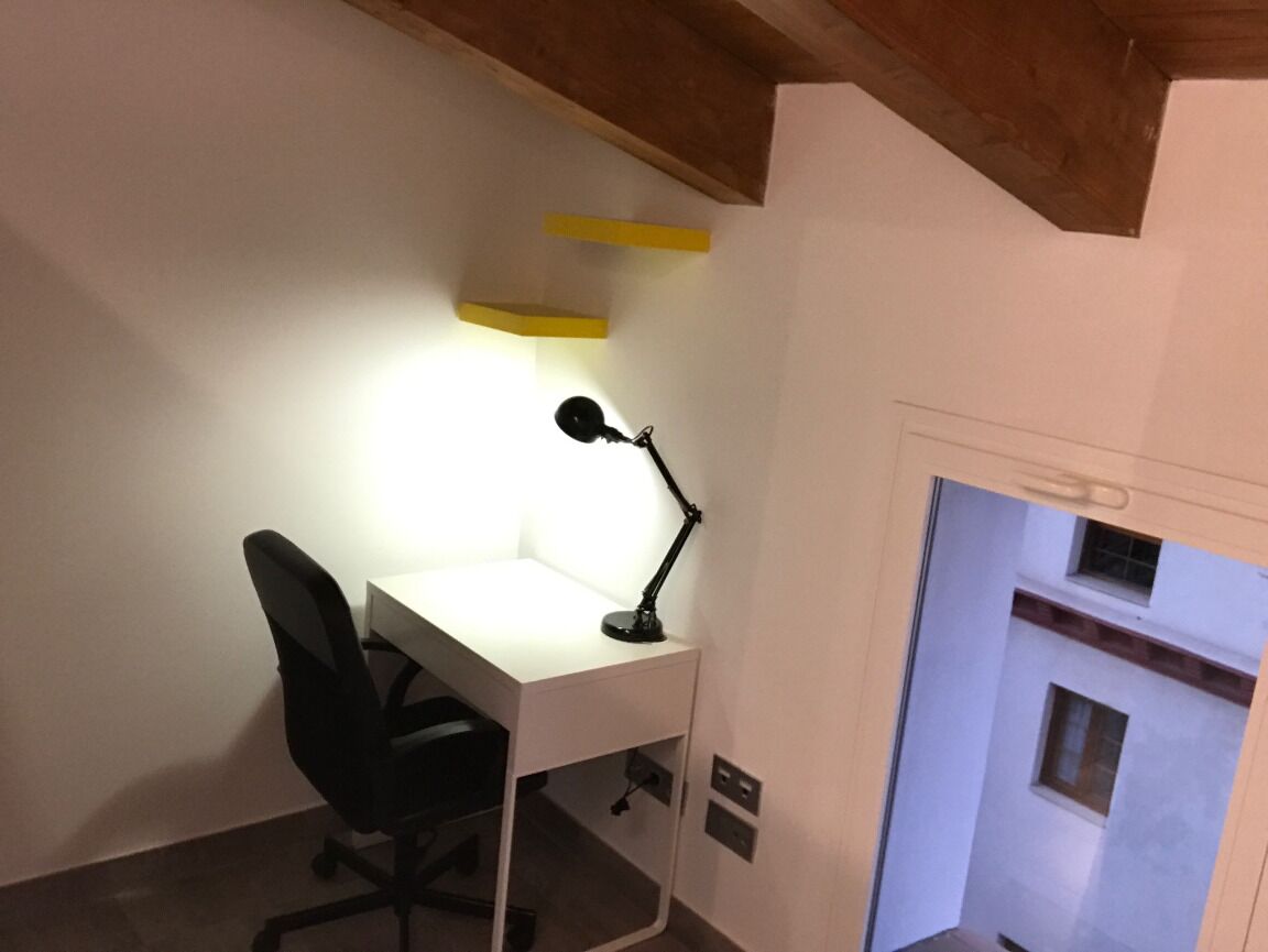 Un pequeño espacio de oficina con un escritorio, una silla y una lámpara de escritorio. Hay una viga de madera en el techo y una pared blanca con un ventanal.