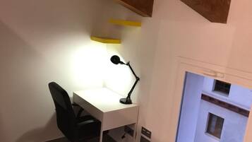 Un pequeño espacio de oficina con un escritorio, una silla y una lámpara de escritorio. Hay una viga de madera en el techo y una pared blanca con un ventanal.