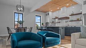 A modern kitchen with white subway tiles, wooden ceiling, and built-in appliances. There are two teal armchairs and a white sofa in the living area. - Luxury Downtown Loft -Walk to Dining-Near Campbell & Ft. Bragg (Lillington)