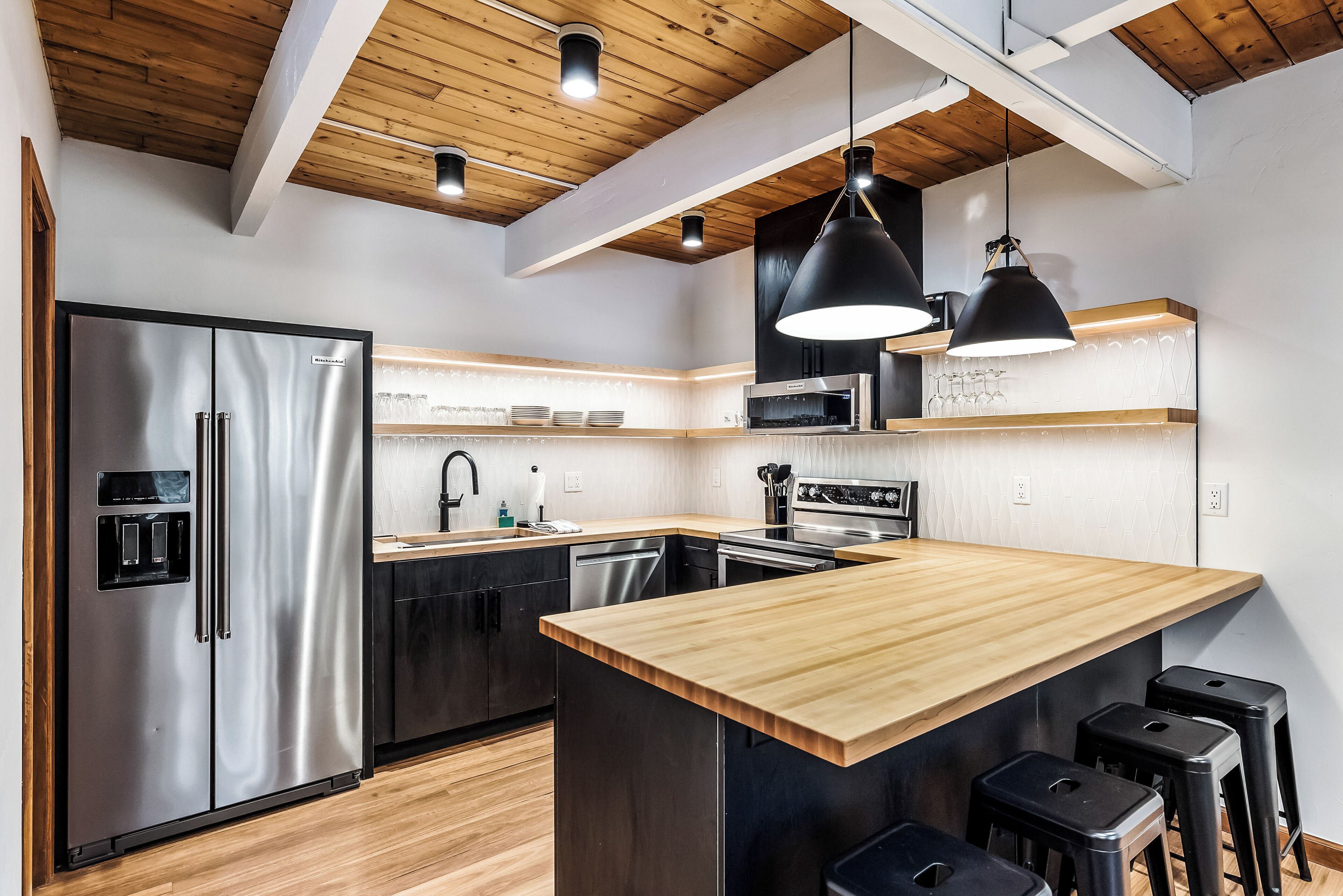 A modern kitchen with a stainless steel refrigerator, wooden countertop, black pendant lights, and a built-in oven.