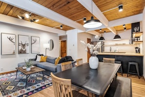 A modern living room with a dining area, wooden ceiling, and a kitchen in the background.