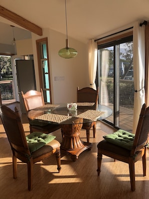 Een eethoek met een ronde glazen tafel, houten stoelen met groene kussens en een hangende groene hanglamp.