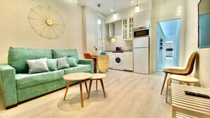 Ein modernes Wohnzimmer mit einem grünen Sofa, zwei kleinen runden Tischen und einer Küchenzeile inklusive Waschmaschine und Schränken.