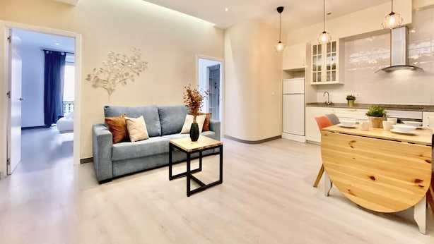 Ein modernes Wohnzimmer mit Sofa, Couchtisch und dekorativer Vase. Angrenzend befindet sich eine Küche mit Holzschränken, einem Kühlschrank sowie ein Essbereich mit Holztisch und Stühlen.