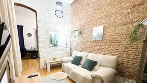 Uma sala de estar acolhedora com parede de tijolos, um sofá branco com almofadas verdes, uma pequena mesa de madeira e uma mesinha lateral redonda.
