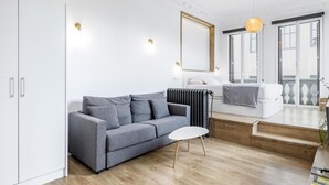 A modern living room with a grey sofa, a small white table, and a radiator. There is a white bed with a grey blanket and a window with a view of the outside. - Home Suite Home I - Studio apartment with terrace in the center (Madrid)
