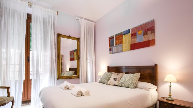 Un lit bien fait, avec des oreillers et des serviettes pliées, une table de chevet en bois avec une lampe, un miroir et un tableau abstrait aux couleurs vives accroché au mur.