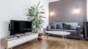 A modern living room with a flat-screen TV on a white cabinet, a grey sofa with cushions, a white coffee table, and a tall green plant. - Home Suite Home III - Studio apartment with large shared terrace (Madrid)