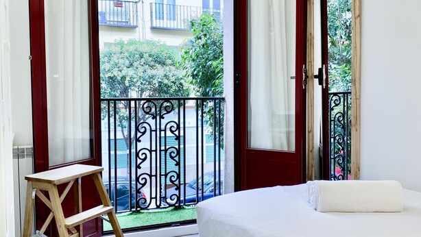 A bedroom with a bed, a wooden ladder, a balcony with a metal railing, and a view of buildings and greenery outside. - Puerta de Toledo VII - 2-Bedroom Apartment (Madrid)