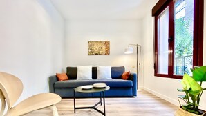 A modern living room with a sofa, a coffee table, and a chair. There is a window with a view of greenery outside. - Puerta de Toledo VII - 2-Bedroom Apartment (Madrid)
