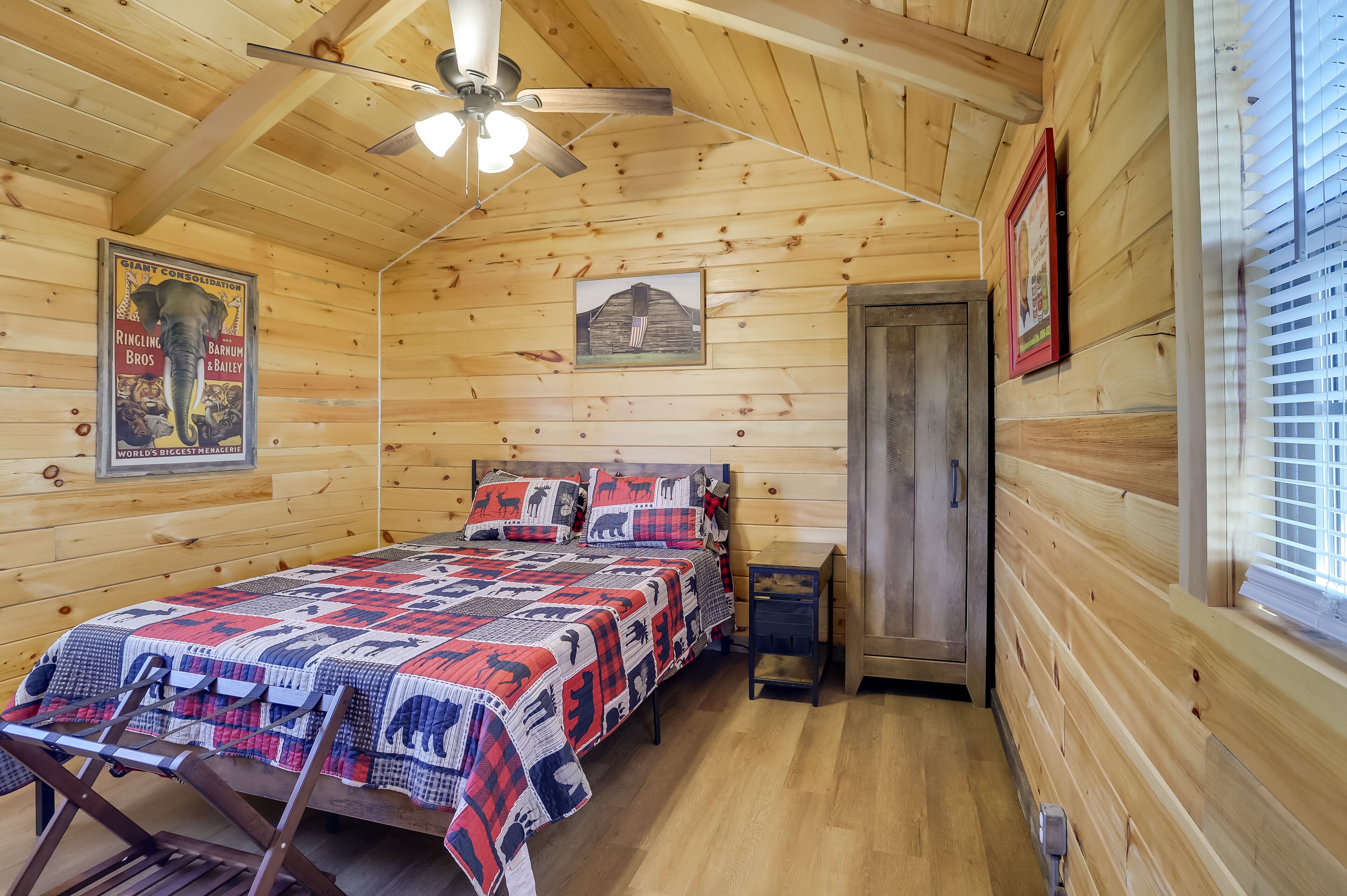 Una habitación tipo cabaña de madera con cama, ventilador de techo, un cuadro enmarcado y una ventana con persianas.