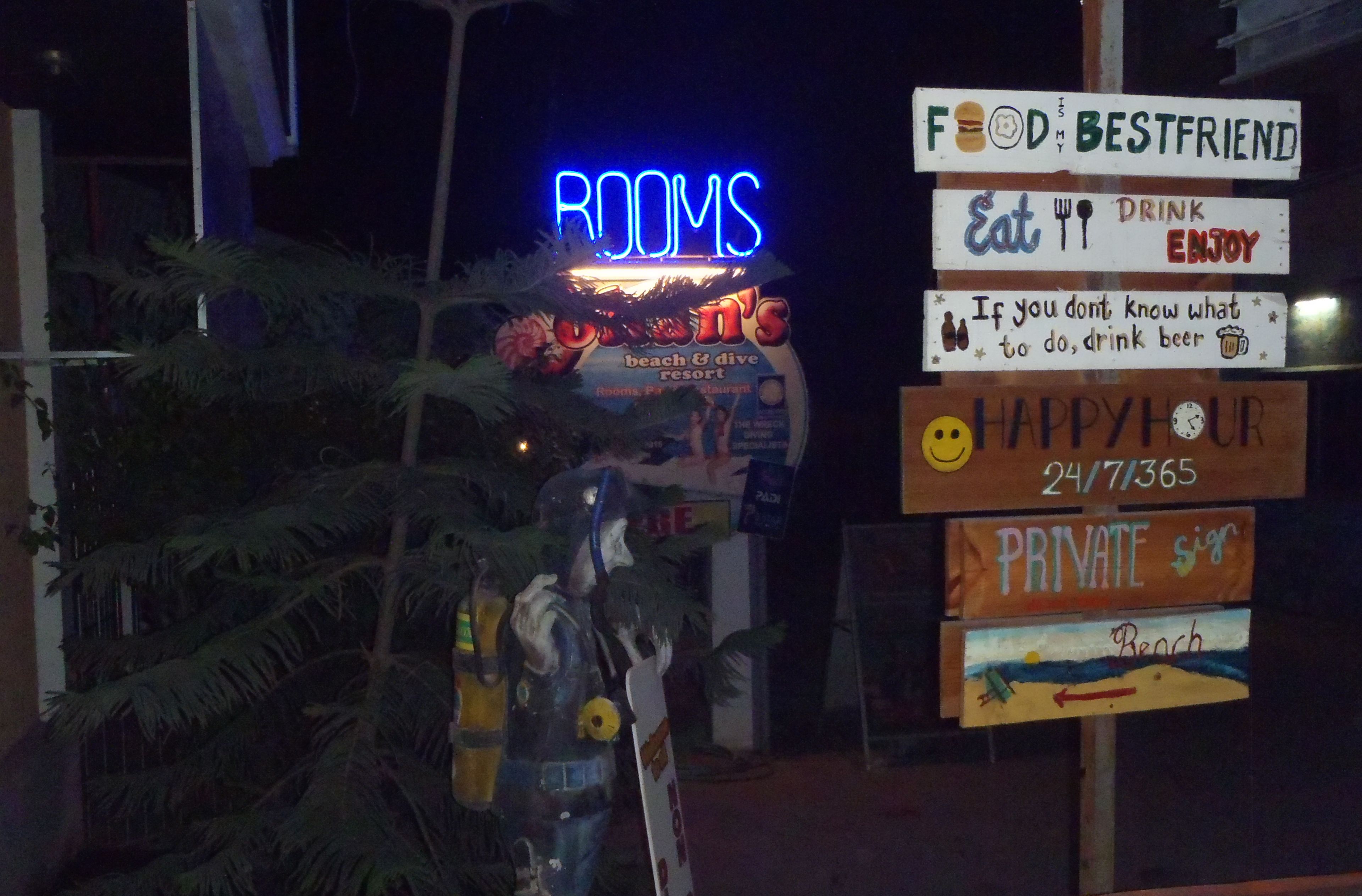 Un cartel con varios letreros que incluyen 'HABITACIONES', 'Food is Bestfriend', 'Comer, beber y disfrutar', 'Si no sabes qué hacer, bebe cerveza', 'Hora feliz las 24 horas, los 7 días de la semana, los 365 días del año', y 'Señal privada'.