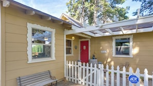 Un bâtiment jaune avec une clôture blanche en bois, une porte rouge et un banc à l’extérieur.
