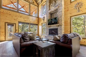 Un intérieur de chalet chaleureux avec une cheminée en pierre, des canapés en cuir et de grandes fenêtres offrant une vue sur la forêt environnante.