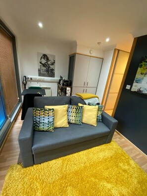 A modern living room with a grey sofa, yellow and patterned cushions, a yellow rug, a kitchen area with cabinets, and a painting on the wall.