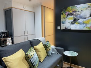 Uma sala de estar moderna com um sofá cinzento, almofadas amarelas e estampadas, uma máquina de café e um quadro na parede.