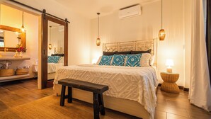 A bedroom with a bed, bedside table, bench, and a bathroom with a sink and mirror.