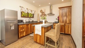 A modern kitchen with wooden cabinets, a stainless steel refrigerator, a built-in microwave, and a bar area with a woven chair.