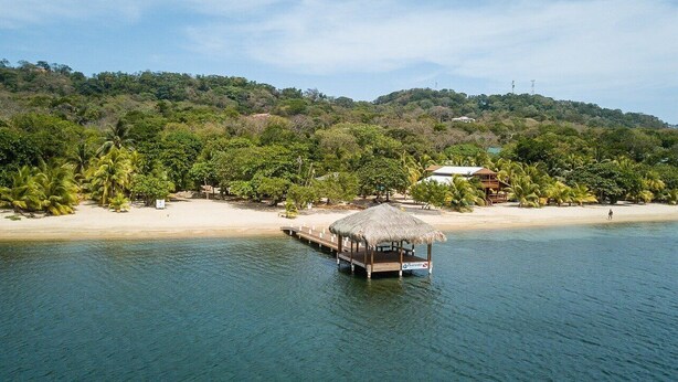 A beach with a thatched-roof structure on stilts in the water, surrounded by palm trees and a hill with buildings in the background.