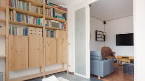 A modern living room with a wooden bookshelf, a grey sofa, a brown armchair, and a flat-screen TV mounted on the wall. - GuestHost - APT with large terrace - Calvairate neighborhood (Milano)
