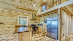 A wooden cabin interior with a kitchen area featuring a large island, stainless steel appliances, and a loft bedroom with a balcony. - Family Retreat in this Gorgeous Log Cabin overlooking the Arkansas Grand Canyon! (Jasper)