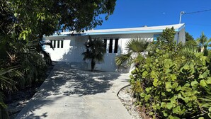 A pathway leading to a white building with a blue roof, surrounded by lush greenery and palm trees. - Bayview Cottage (Staniel Cay)
