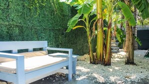 Ein weißes Bett mit einer beigen Matratze in einem Garten mit Hecke und Bananenpflanzen.