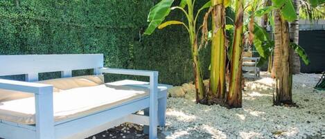A white bed with a beige mattress in a garden setting with a hedge and banana plants.