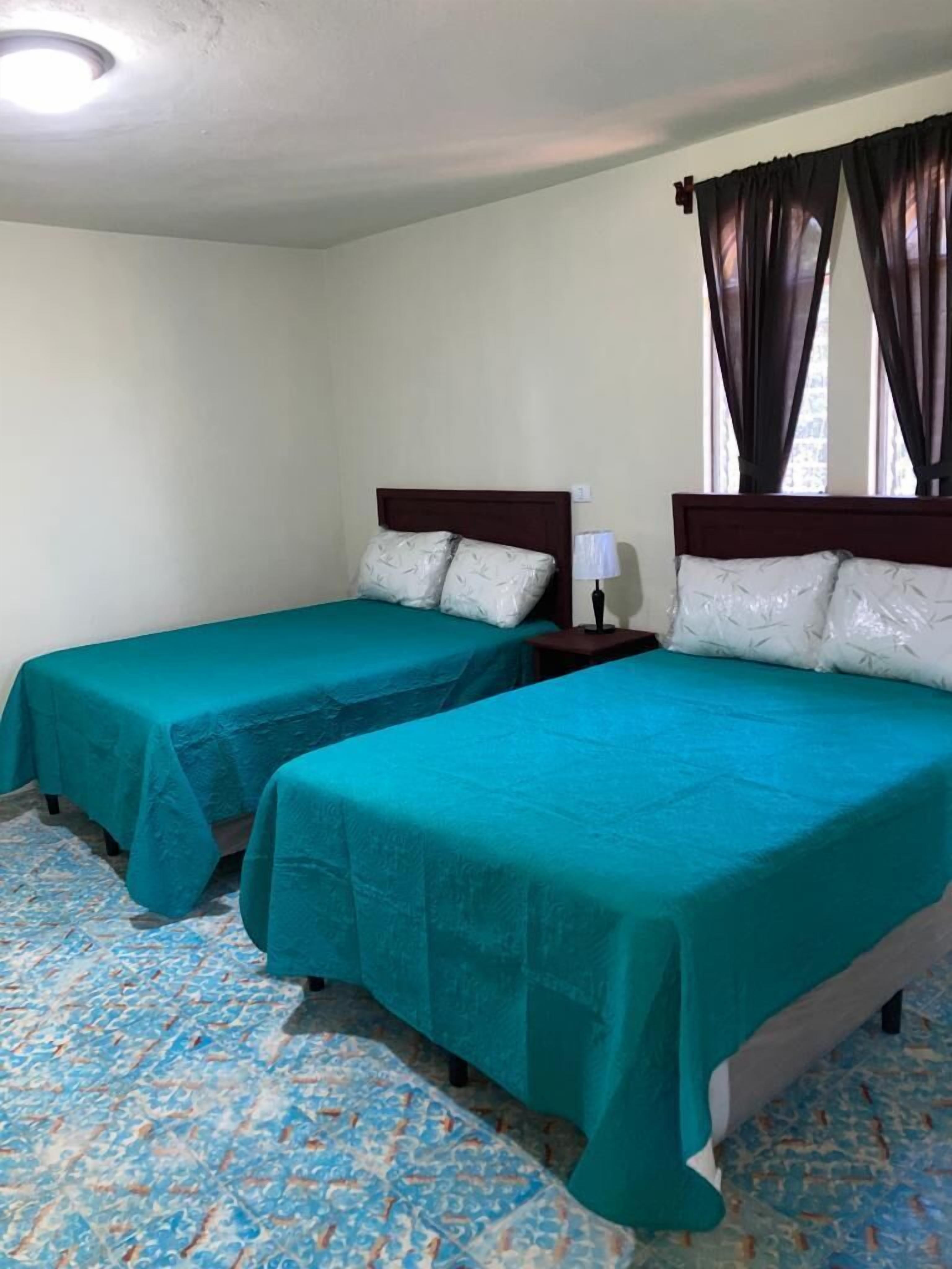 A hotel room with two beds, each with a wooden headboard and teal bedspreads. There is a nightstand with a lamp on one side and a window with dark curtains on the other.