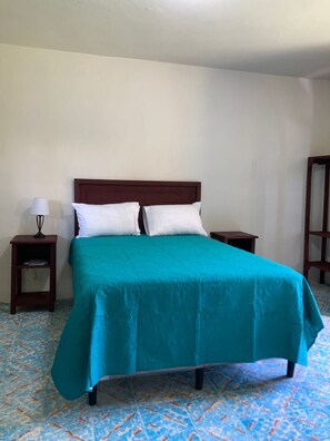 Een enkel bed met een blauwgroene sprei en twee witte kussens, zijkanten met nachtkastjes en lampen, in een kamer met vloertegels met een patroon.
