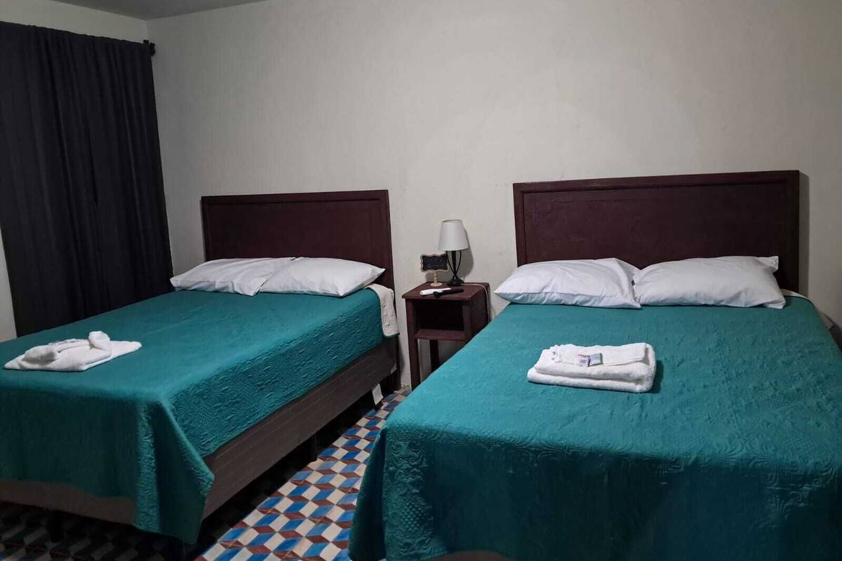 Hotellihuone, jossa on kaksi erillistä sänkyä. Jokaisessa sängyssä on turkoosi sängynpeite ja valkoiset tyynyt. Huoneessa on myös pieni yöpöytä, jossa on lamppu, sekä ruutukuviollinen lattia.