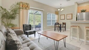 A modern living room with a sofa, a dining table, and chairs. There is a kitchen area with a refrigerator and a window.